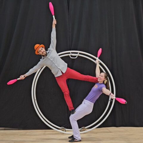 A man and woman pose balanced on each other inside a German wheel. Both are holding bowling pins.